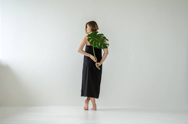 Young European woman in a black long dress, standing with her back to viewer, holding a large monstera leaf in her hands on a white light room background - Foto, Imagem