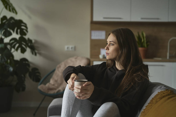 Biała smutna kobieta siedząca na kanapie z filiżanką kawy - Zdjęcie, obraz