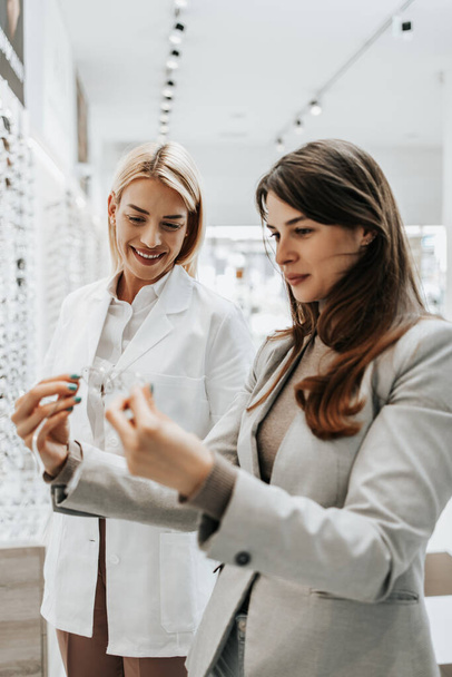 Beautiful and fashionable woman choosing eyeglasses frame in modern optical store. Female seller specialist helps her to make right decision. - 写真・画像