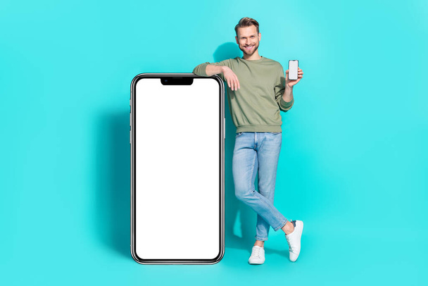 Photo of cheerful man hold telephone show screen empty space wear pullover jeans isolated blue color background. - Foto, Imagen