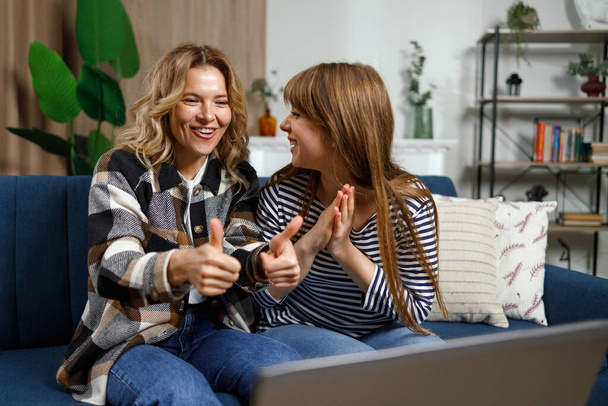 Cheerful mature woman shows thumbs up while sitting with her adult daughter on the couch in front of a laptop talking on a video call or watching video - Photo, Image