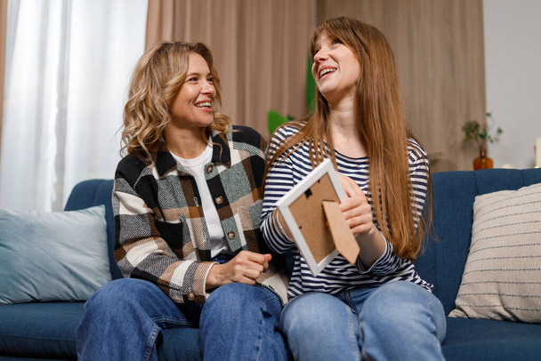 Adult daughter and her mature mother looking at photo in frame while sitting on the sofa in home. Family, generation and people concept  - 写真・画像