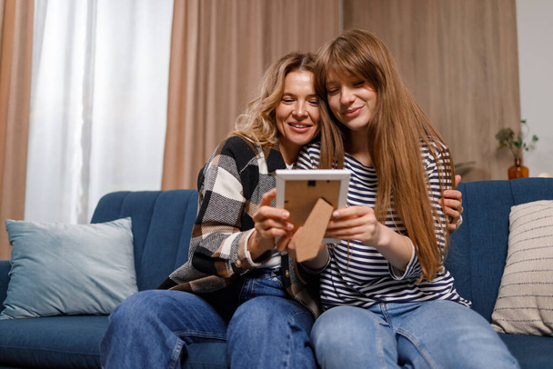 Glückliche Familienmutter und Tochter sitzen auf dem Sofa und betrachten das Rahmenfoto zu Hause - Foto, Bild