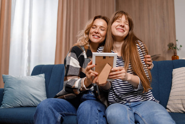 Mãe e filha sentados no sofá na sala de estar abraçando uns aos outros olhando para uma foto antiga em uma moldura - Foto, Imagem