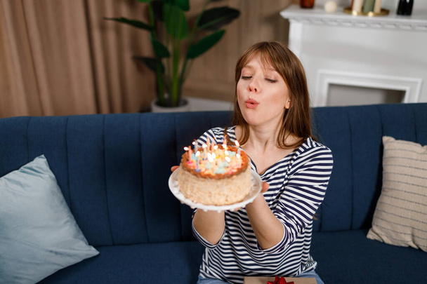 Giovane donna caucasica che fa desiderio di compleanno e soffia candela sulla torta seduta con gli occhi chiusi sul divano a casa - Foto, immagini