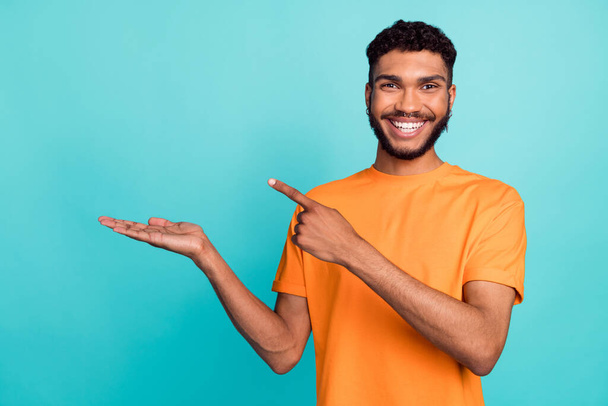 Photo of pretty positive guy wear orange t-shirt pointing finger holding arm empty space isolated teal color background. - Foto, Bild