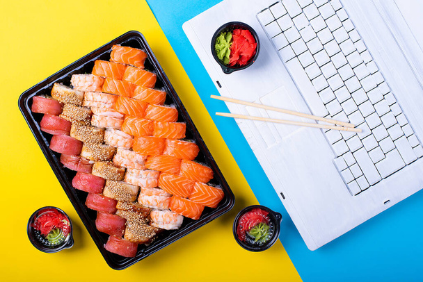 Semmeln, Sushi und Laptop im Büro auf blauem und gelbem Hintergrund. Japanisches Mittagessen am Arbeitsplatz. Lachs und Wasabi Sojasauce - Foto, Bild