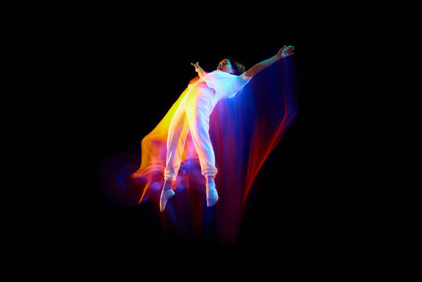 Levitation. One flying, jumping dancer or gymnast performing tricks in the air over black background with mixed neon glowing rays. Fantasy, cyberpunk, sport, fashion - Photo, Image