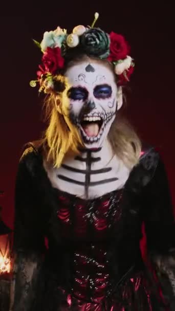 Vertikales Video: Horrorgöttin des Todes schreit im Kostüm mit Totenkopf-Körperkunst, trägt eine Blumenkrone und schreit wie der heilige Weihnachtsmann, um dios de los muertos zu feiern. Frau am Übergang - Filmmaterial, Video