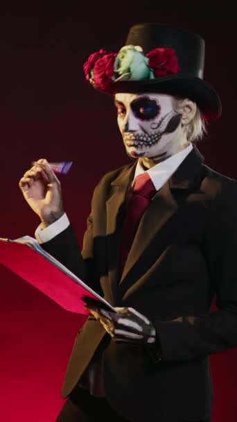 Vertical video: Creepy horror woman taking notes on clipboard papers, looking like santa muerte with day of the dead make up on mexican holiday celebration. Lady of death traditional costume with suit - Footage, Video