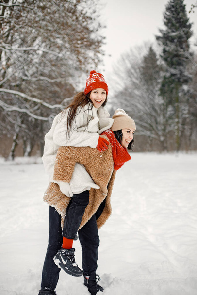 Joyful girl and her beautiful mother spending time together in the winter park. Woman wearing warm jacket and red sweater. Mother carrying her daughter piggyback. - Foto, imagen
