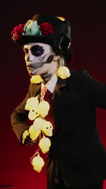 Vertikální video: Děsivá žena v kostýmu smrti tančí ve studiu, poslouchá hudbu na sluchátkách a dělá taneční pohyby. Vypadáš jako svatá mexická bohyně na oslavu dios de los muertos - Záběry, video