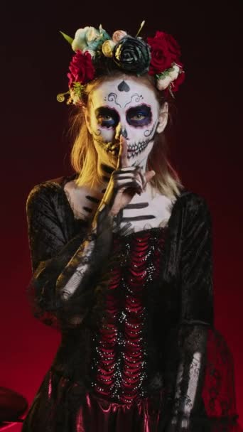 Vertical video: Young scary woman doing hush gesture to keep secret, wearing santa muerte costume and showing slience mute and secrecy sign with finger over lips. Holy dios de los muertos holiday. - Footage, Video