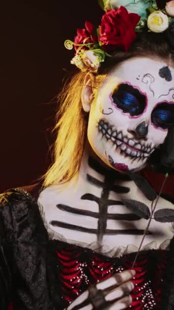Vertical video: Beautiful model wearing death costume and flowers wreath over studio background, acting like holy santa muerte. La cavalera catrina lady of dead with skull body art, mexican culture - Footage, Video