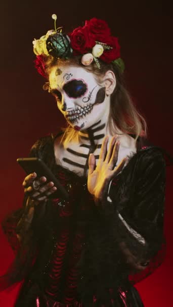 Vertikální video: Okouzlující děsivá žena pomocí smartphonu procházet internetové stránky přes černé pozadí. Santa Muerte model zobrazující horor bohyně smrti na mexické sváteční oslavy ve studiu. - Záběry, video
