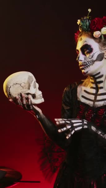 Vertical video: Creepy dead woman holding holy skull in studio, acting scary and horror to celebrate mexican halloween day. Одинокие богини в фестивальном костюме с боди-артом, похожие на дам - Кадры, видео