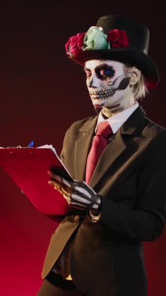 Vertical video: Dios de los muertos model taking notes on papers, looking like santa muerte with day of the dead make up on mexican holiday celebration. Lady of death traditional costume with suit and - Footage, Video
