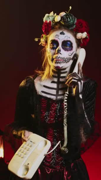 Vertical video: Spooky woman answering landline phone with cord, talking on office telephone while she has skull make up and body art. Portraying santa muerte lady of death on dios de los muertos - Footage, Video