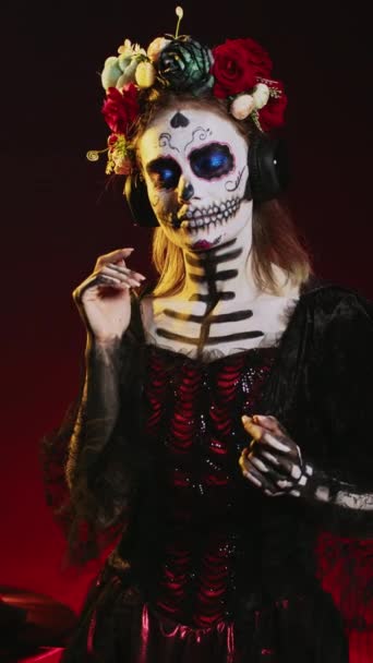 Вертикальное видео: Богиня мертвых, наслаждающаяся музыкой на наушниках, носящая макияж черепа и костюм Санта-муэрте в студии. Человек слушает песню на аудиогарнитуре, празднуя день мертвых - Кадры, видео
