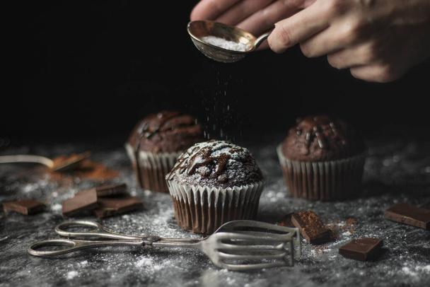 Close up of chocolate muffins with icing and hands decorating them with sugar, atmospheric dark still life - Photo, image