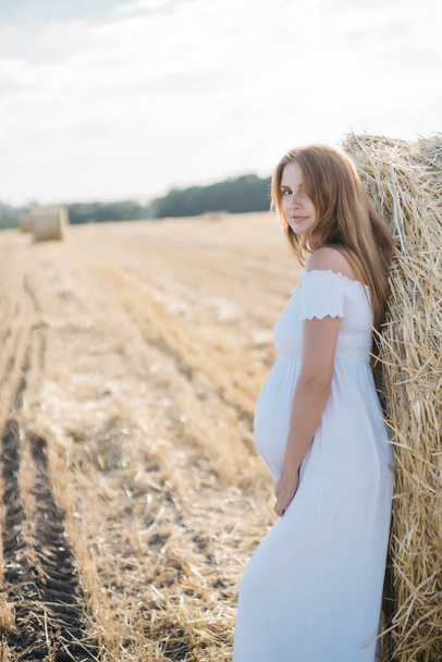bella bruna incinta in un vestito bianco vicino ai pagliai nel campo al tramonto. Foto di alta qualità - Foto, immagini