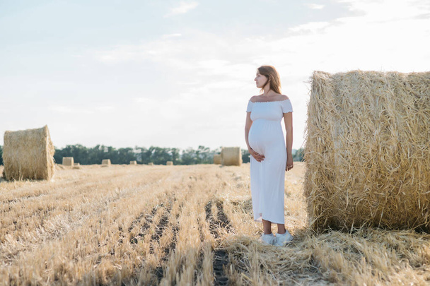 bela morena grávida em um vestido branco perto dos palheiros no campo ao pôr do sol. Foto de alta qualidade - Foto, Imagem