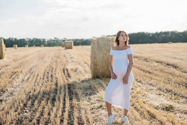 beautiful pregnant brunette in a white dress near the haystacks in the field at sunset. High quality photo - Foto, imagen