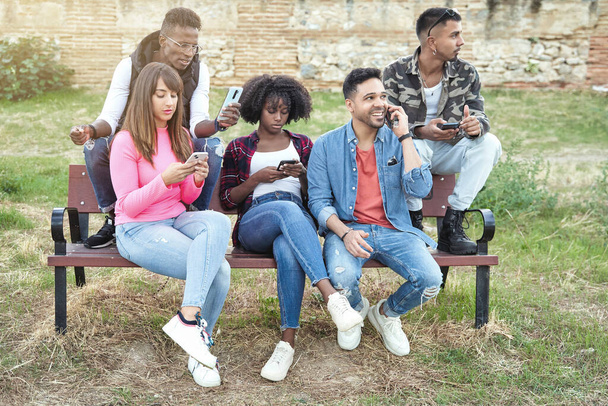 Multiethnische Freunde benutzen ihre Mobiltelefone, während sie im Freien auf einer Bank sitzen. Technologie und Freundschaftskonzept. - Foto, Bild