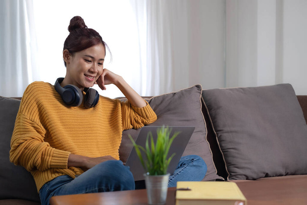 Candid of asian woman using laptop for watching online movie stream mobile device on sofa at living room. Young happy girl enjoying with entertainment media online on weekend.. - Foto, Imagen