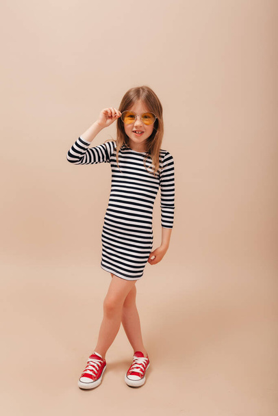 Full-lenght photo of stylish little girl with happy smile wearing striped dress and red sneakers touching round orange glasses and standing over beige background - Valokuva, kuva