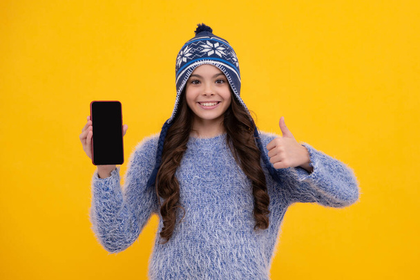 Kid teen child in winter knitted hat showing blank screen mobile phone, mock up copy space. Happy face, positive and smiling emotions of teenager girl - Fotografie, Obrázek