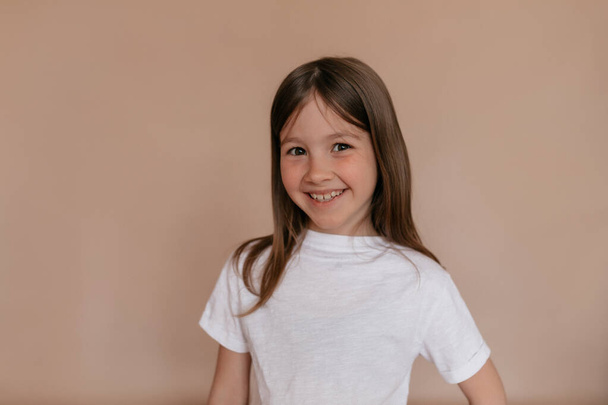 Studyjne ujęcie małej europejskiej stylowej dziewczyny z piegami i cudownym uśmiechem w białej koszulce uśmiechniętej na odizolowanym beżowym tle  - Zdjęcie, obraz