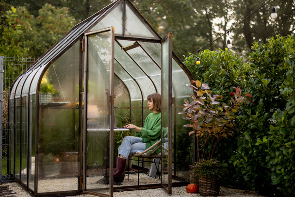 Die junge Frau arbeitet am Laptop, während sie am runden Tisch im Glashaus sitzt und im Hinterhof Pflanzen und Blumen präsentiert. Von zu Hause aus in gemütlicher Atmosphäre an der Natur arbeiten - Foto, Bild