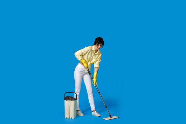 charming housewife girl in rubber gloves washing floor with mop on blue background. Full length - Foto, afbeelding