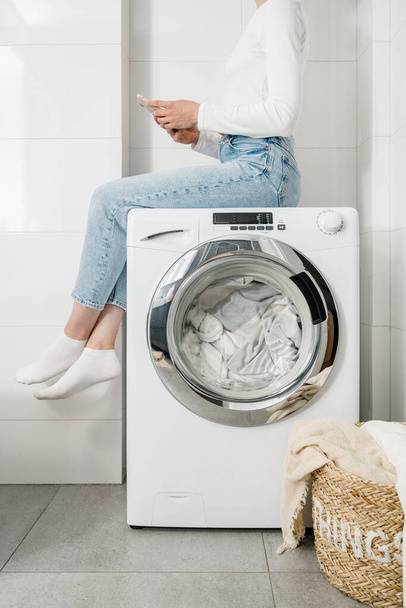 vrouw huisvrouw zitten met mobiele telefoon op automatische wasmachine met beddengoed binnen en gesloten deur, rieten mand met kleren op de vloer in de badkamer, bijgesneden schot - Foto, afbeelding