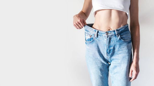 脂肪吸引、ケトやパレオ食事の後の結果を示すオーバーサイズのジーンズでスリムな女性の作物のショット。減量の概念。コピースペースの背景に立って完璧な腹筋を持つスポーティ女性 - 写真・画像
