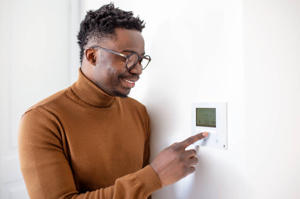 Smiling African American man using modern smart home system, controller on wall, positive young man switching temperature on thermostat or activating security alarm in apartment - Foto, afbeelding