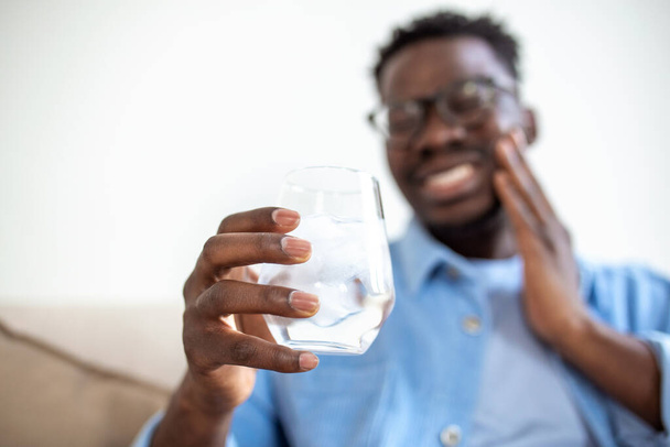 Young man with sensitive teeth and hand holding glass of cold water with ice. Healthcare concept. man drinking cold drink, glass full of ice cubes and feels toothache, pain - Photo, image