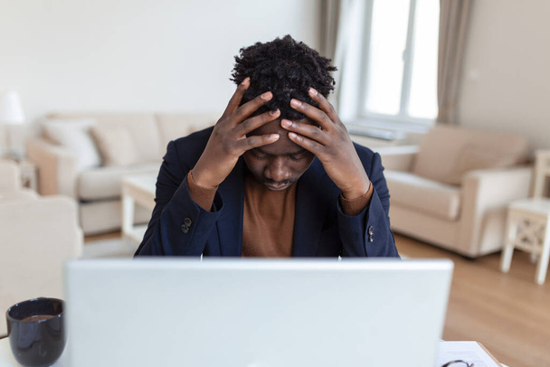 Stressaantunut väsynyt afroamerikkalainen mies koskettaa temppeleitä, kärsii päänsärystä pitkän työpäivän jälkeen, ylityöllistetty liikemies istuu pöydällä, tunne huonovointisuus - Valokuva, kuva
