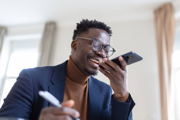 職場からの電話で話している間、アフリカのビジネスマンはノートをメモ帳に書いています。音声認識の概念です。手のないコミュニケーション. - 写真・画像