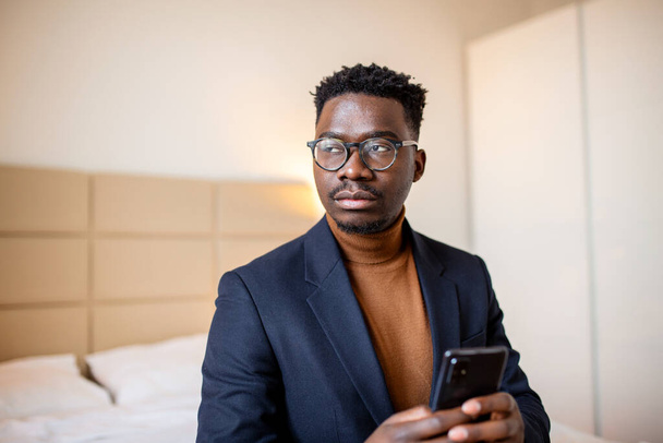 Hombre de negocios africano con smartphone en una habitación de hotel. Un hombre guapo parado en la ventana, mandando mensajes de texto. De cerca.. - Foto, Imagen