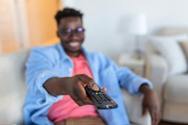Захоплена афроамериканська людина вказує телевізійний контролер для перемикання камер Перегляд телевізора. Телевізійна реклама. Вибірковий фокус
 - Фото, зображення
