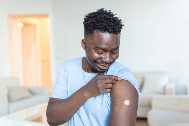 Портрет человека, улыбающегося после получения вакцины. Африканский мужчина, держащий рукав рубашки и показывающий руку с бинтом после вакцинации. - Фото, изображение