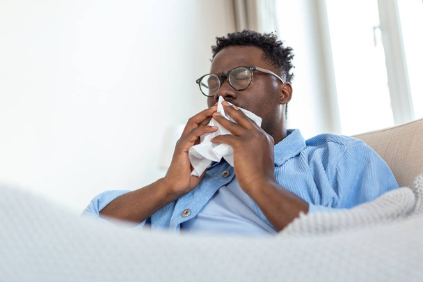 Kranke afrikanische junge Frau, die mit einer Decke bedeckt ist, bekommt Fieber, wenn sie erkältet niest, sitzt auf dem Sofa, krankes allergisches schwarzes Mädchen mit Allergiesymptomen, die zu Hause husten, GrippekonzeptKranker afrikanischer junger Mann, der mit einer Decke bedeckt ist - Foto, Bild