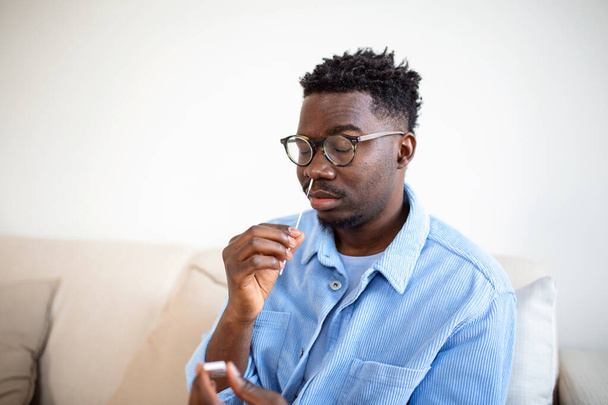 Афро-американец использует ватный тампон во время проведения ПЦР у себя дома. человек с помощью коронавируса быстрого диагностического теста. Молодой человек дома использует мазок из носа для COVID-19. - Фото, изображение