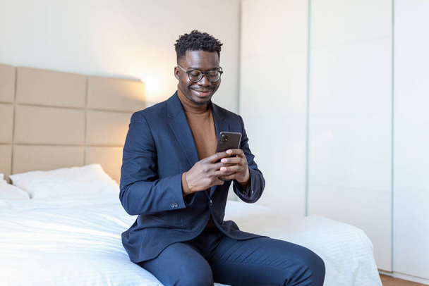 Африканский бизнесмен со смартфоном в номере отеля. Красивый мужчина стоит у окна и пишет смс. Закрыть. - Фото, изображение