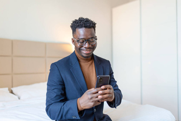 Homme d'affaires africain avec smartphone dans une chambre d'hôtel. Un bel homme debout à la fenêtre, envoyant des textos. Gros plan. - Photo, image