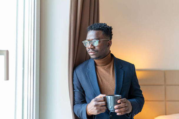 Бізнесмен стоїть біля вікна готелю і п'є каву. Духовний молодий африканський бізнесмен стоїть біля вікна з чашкою кави і дивиться назовні.. - Фото, зображення