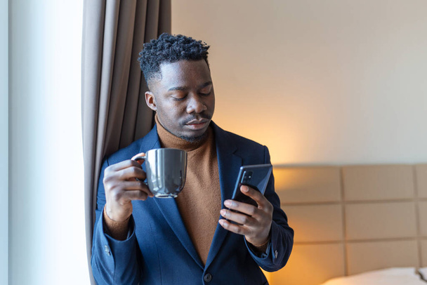 Επιχειρηματίας στέκεται δίπλα στο παράθυρο στο δωμάτιο του ξενοδοχείου πίνοντας καφέ. Ευγενικός νεαρός Αφρικανός επιχειρηματίας στέκεται δίπλα στο παράθυρο με ένα φλιτζάνι καφέ και κοιτάζοντας το κινητό του τηλέφωνο. - Φωτογραφία, εικόνα