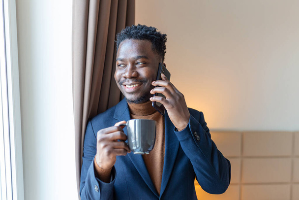 Empresario de pie junto a la ventana en la habitación del hotel bebiendo café. Pensativo joven empresario africano de pie junto a la ventana con una taza de café y mirando su teléfono móvil. - Foto, imagen